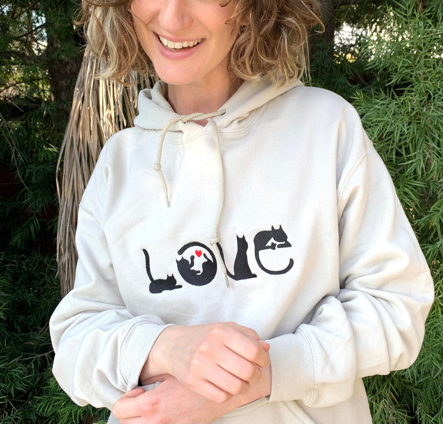 Smiling young woman wearing sand colored hoodie with image of black cats spelling out the word LOVE with a red heart inside the o.