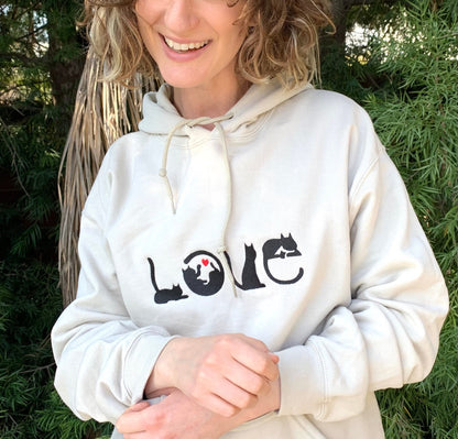 Smiling young woman wearing sand colored hoodie with image of black cats spelling out the word LOVE with a red heart inside the o.